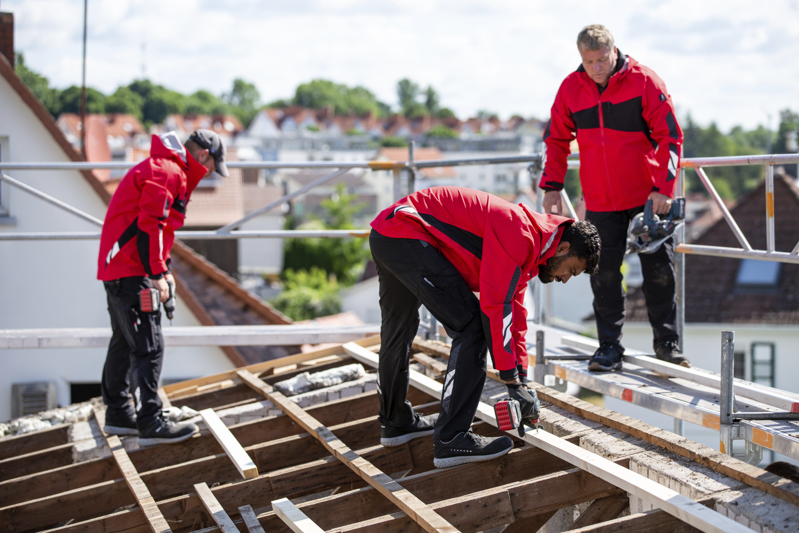 Drei Arbeiter in schwarzen Arbeitshosen und roten Arbeitsjacken die auf dem Dach Laten festschrauben.